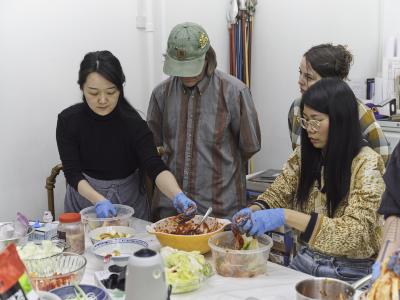 Four guests stand around a table, as two, with gloved hands massage a red sauce into cabbage. The other two with arms behind their back pay close attention.