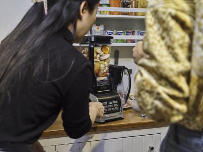Two guests, with their backs to the camera, stand around a blender full of garlic, onion, and bell peppers.