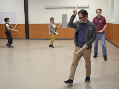 Participants walk in a circle wielding wooden sticks ready to hit them together. The leader appears meditative while the others follow their steps. 