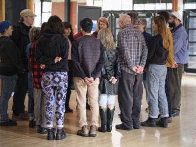 A group of people gather in a circle facing each other. They stand close together with hands clasped either in front or behind them.