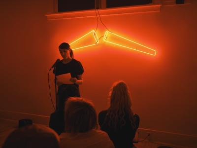A presenter, dressed in black, holds a single piece of paper and reads aloud. They wear straight hair in a low ponytail, and hold the paper by their waist. They stand in front of neon red lights shaped like cones.
