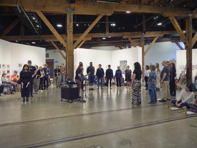 Some participants walk around the venue banging wooden percussion sticks while others stand in a semicircle. The audience watches this movement.