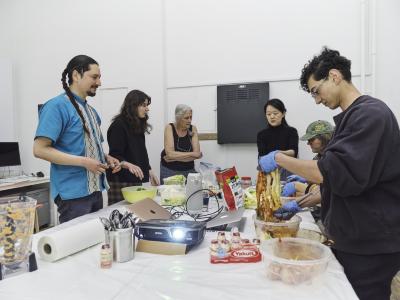 Guests gather around a plastic lined table. Half of the group wears gloves while massaging cabbage with kimchi sauce. The others watch keenly.