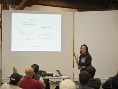A presenter speaks in front of a seated audience. They hold a balingbing, resembling the diagram projected behind them.