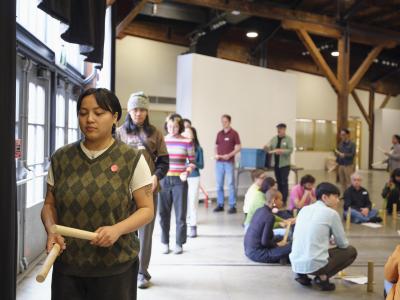 A group of participants walk banging rounded wooden sticks together. They circle another group which is seated on the floor.