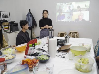Guests sit around a table covered in chopped produce and appliances. An artist stands up with hands clasped motioning towards a blender. Projected behind them is a Zoom meeting showing participants tuning in.