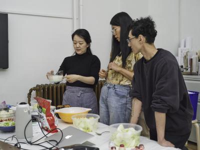 Three guests stand around a table line with food, utensils, and electronics. One guest holds and points to a milky liquid while the others observe.