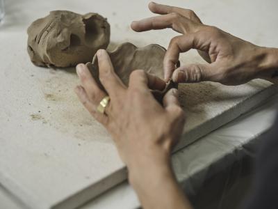 Two hands pinch the side of a small concave clay sculpture. They use only a pointer finger and thumb on each hand extending the others so that they are out of the way.