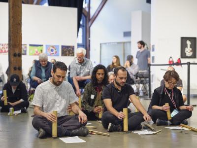A group of participants sit on the ground stamping tongatongs on the floor. They concentrate on the sheet of music laying before them. Audience members are seated behind them in chairs.