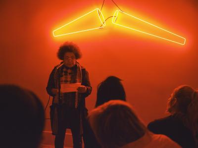 A presenter, dressed in a black coat and tie-dye shirt, holds onto a page of paper with two hands. They wear curly hair above their shoulders. They stand in front of orange lights which fill the room in their hue. 