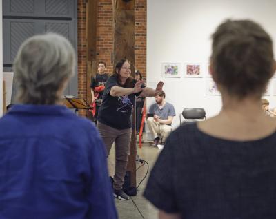 Two participants, with their backs facing the camera, give their attention to another participant who extends their arms in front of their chest. An audience member films the scene.
