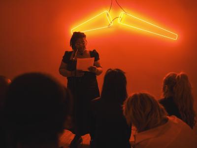 A presenter, dressed in a black dress, holds a piece of paper near their chest. They wear twisted hair to the length of their shoulders and a necklace with a black charm. They stand in front of red neon lights shaped like cones.
