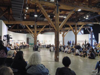 A wide shot of the performance space showing a large gathering of people. Some are seated on the ground with instruments in front of them, while others are seated in chairs. A speaks into a make at the center of the gathering with cameras pointed at them from across the room. 