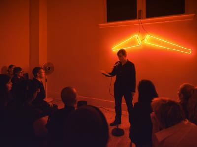 A presenter, dressed in a black button up, holds a piece of paper in their right hand, with the left one resting at their side. They wear a short hairstyle, with straight hair falling right above their ears. The orange glow of neon lights shine on faces in the audience. 