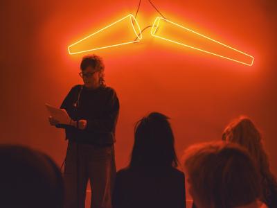 A presenter, dressed in a black sweater and wearing reading glasses, holds two sheets of paper to their right side. Their hair is fashioned into a curly mullet reaching their shoulders. They stand in front of two cone-shaped neon lights and read out to the audience.