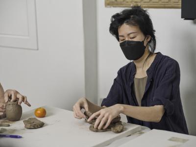 A participant in a navy button up top collects flattened pieces of clay together in front of them. To their right, sits a tangerine and the hands of another guest working away.