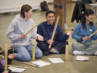 Three participants smile as they play the balingbing. They sit on the ground with sheet music and other instruments in front of them.