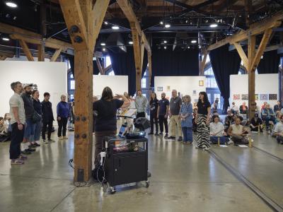 A group of participants stand in a half circle with their mouths widened paying attention to a conductor with their arms extended. Seated on the floor next to them are performers playing balingbings. Behind these performers you can spot audience members watching the event.
