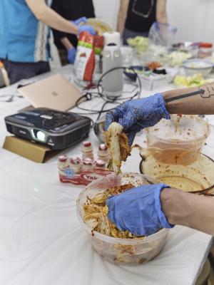 Gloved hands reach onto a tupperware of kimchi. A projector glows, directing its light out of the frame. Behind bottles of Yakult, cables, and a laptop, are more gloved hands transferring kimchi from one bowl to another. 