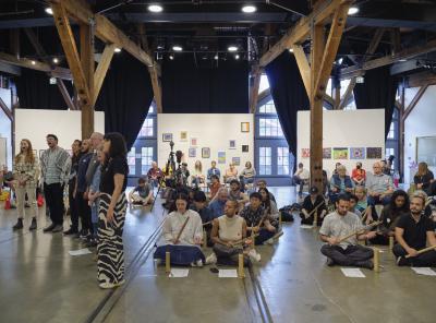 Rows of participants are seated on the floor playing balingbings. Next to them stand a group of participants singing. Behind them, audience members watch the event.