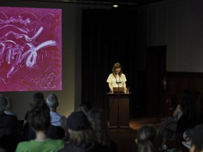 A woman with brown shaggy hair stands at a wood lectern under a spotlight and reads from her laptop. She wears a white t-shirt. To her right, a pink abstract image is projected on a screen. Seated audience members can be seen from behind.