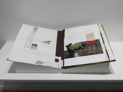 A mixed-media artist book lies open on an angular plinth. The spread is a collage with  stitching, tape, handwritten text, and drawing.