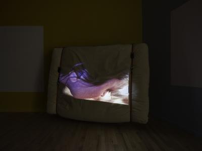 A video is projected on a mattress strapped to a wooden pallet. The still shows Sheilah ReStack’s face in profile.