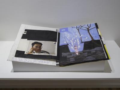 A mixed-media artist book with collage, drawing, and handwritten text lies open on an angular plinth.