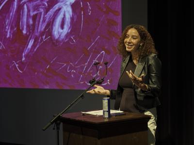 A woman with curly copper hair stands at a wood lectern, smiling while casually gesturing with her hands. She wears green earrings, a leather jacket over a black top, and white pants. A stack of paper and a can of sparkling water are arranged on the surface in front of her. In the background, a pink and red abstract print is projected on a screen.