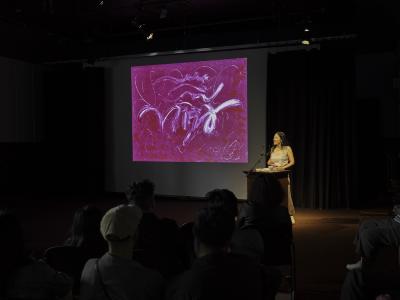 A woman with long middle-parted black hair stands at a wood lectern next to a projector screen displaying a pink abstract print. She wears a grey tank top and taupe skirt. Seated audience members can be seen from behind.