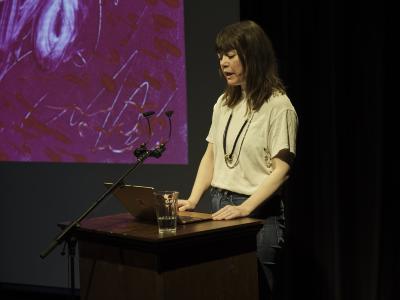 A woman with brown shaggy hair stands at a wood lectern, smiling. Her fingers hover on her Macbook keyboard as she gazes out into the audience. She wears a white t-shirt, layered necklaces, and blue jeans. The corner of a pink abstract image can be seen projected on an adjacent screen.