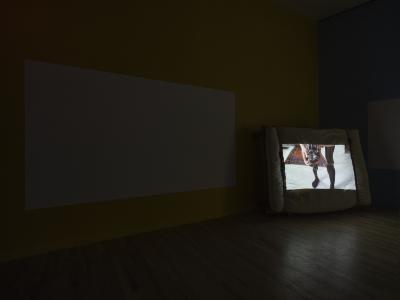 A video is projected on a mattress strapped to a wooden pallet. The still shows a person holding an analogue camera.