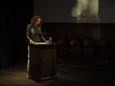 A woman with curly copper hair stands at a wood lectern, reading from loose sheets of paper. She gazes towards her notes, which are arranged next to a blue can of soda water. She wears a leather jacket over a black top, white pants, and sandals. Behind her, light filters through a window out of frame and casts diffused patterns across the wall.