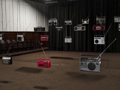 Radios hang in rows from the ceiling in a carpeted room. Three amongst the many silver and black radios are red. Lights shining from above project black shadows looming below. 