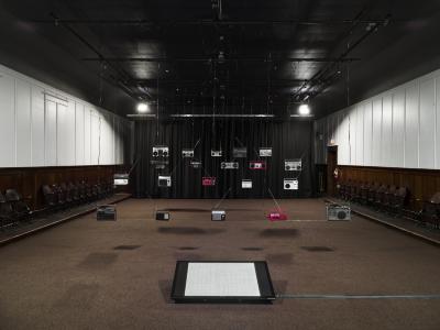 Twenty radios hang from wires, getting closer to the ceiling as they reach the back of the room. A television, propped up from the floor displays a musical score sorted into 300 cells.