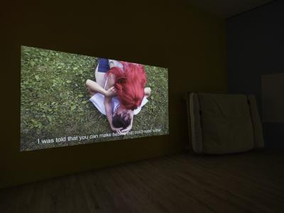 A video is projected on a wall. The still shows Dani and Sheilah Restack making out in the grass. Sheilah straddles Dani and wears a cherry red wig. White text runs across the bottom of the video that reads, I was told that you can make babies that don’t need water.