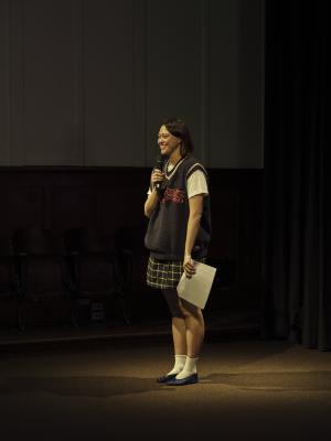 A woman with a brown bob haircut stands under a spotlight and smiles while holding a microphone up to her mouth in one hand, and speaking notes in the other. She wears an oversized navy knit sweater vest over a white t-shirt, a plaid mini skirt, and white socks with blue satin flats.