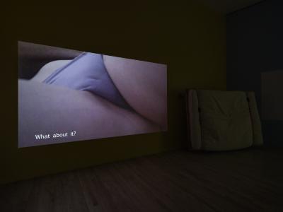 A video is projected on a wall. The still shows a close-up of a woman’s crotch from below. She lies down and is wearing periwinkle bikini underwear. White text runs across the bottom of the video that reads, What about it?