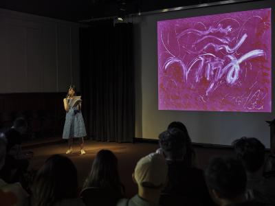 A woman wearing a blue short-sleeved patterned dress and white sneakers reads from a stack of papers into a cordless mic. She is lit by a spotlight and stands adjacent to a pink abstract print projected on a screen. 