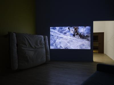 A video is projected on a blue wall. The still shows a close-up of a sewing machine foot as it punctures a paper surface with stitches.