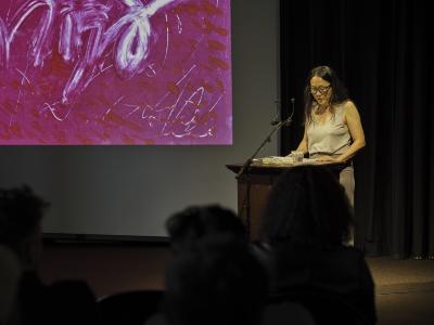 A woman with long middle-parted black hair reads at a wood lectern under a spotlight. She wears black framed glasses, a grey tank top, and a taupe skirt. To her right, a pink abstract print is projected on a screen.