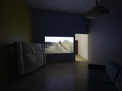 A video is projected on a blue wall. The still shows a person walking outdoors down a path lined with tall grass.