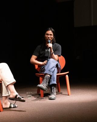 A man sits in a red chair and speaks into a microphone. He has black mid-length hair, and wears glasses, a black t-shirt with white writing, cuffed blue jeans, and black combat boots. A glass of water is positioned on the floor next to his chair. 