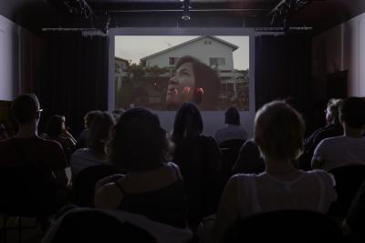 An auditorium full of people sit facing a screen that shows a Filipino woman standing in front of a house, gazing up towards the sky. The image is overlaid with a hand with glowing fingertips.