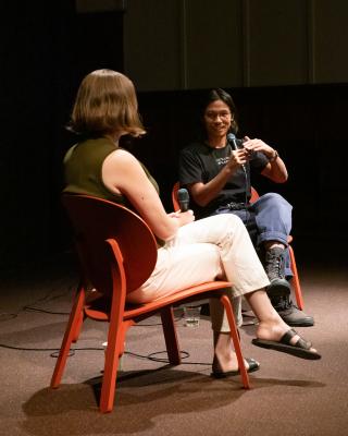 Two people sit facing each other in red chairs under a spotlight. They are in conversation, and each holds a microphone. A man with black hair and glasses speaks enthusiastically to a woman with a blonde bob, who is facing away from the camera.