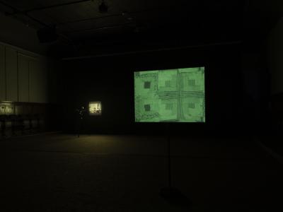 Two photosensitive screens are arranged in a dark room that display green hued photographs: one of a candid family photograph and the other of a patchwork textile.