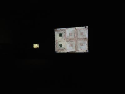 Images are projected onto two screens in a darkened room. One screen, which is further away from the camera, shows a candid family photograph. The other screen shows a patchwork textile.