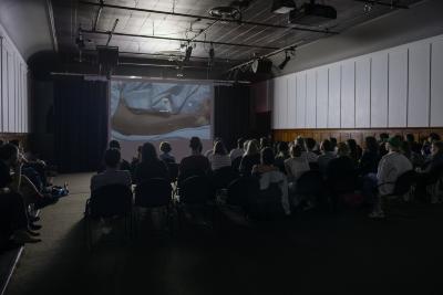 An auditorium full of people sit facing a screen that shows an image of an arm wrapped in blue sheets overlaid with an opaque image of bubbles floating on the surface of the water. The words nowhere near are written in small white text at the centre of the screen. 