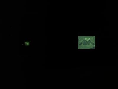 Two photosensitive screens are arranged in a dark room that display green hued photographs: one of the artist’s grandmother and the other of a jacket laid on a table and overlaid with an abstract pattern.