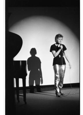 A person wearing kitten heels and shiny pants stands with a wired microphone to their mouth. Behind them is a large flat screen where a spotlight creates a crescent shape around their body. A long shadow is cast behind them, echoing their silhouette.
